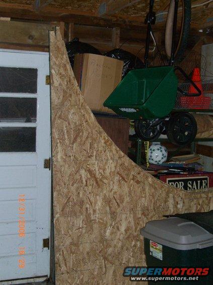 Garage scene with a wooden ramp against a door, storage shelves with a green wheelbarrow, and a trash bin