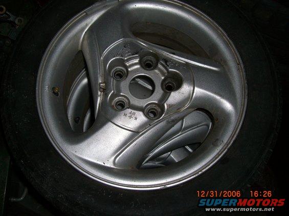Close-up of a silver alloy car wheel with five lug nut holes, resting on a tire. Some markings are visible on the wheel