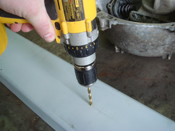 hand holding yellow drill, drilling into a light gray metal bar. Metal casing in background