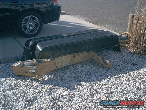 trailer hitch and part of a trailer frame on gravel, with a black car in the background