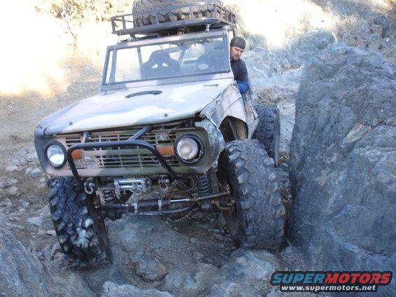 Modified off-road SUV with large tires and a roof rack, driving over rocks. Driver visible
