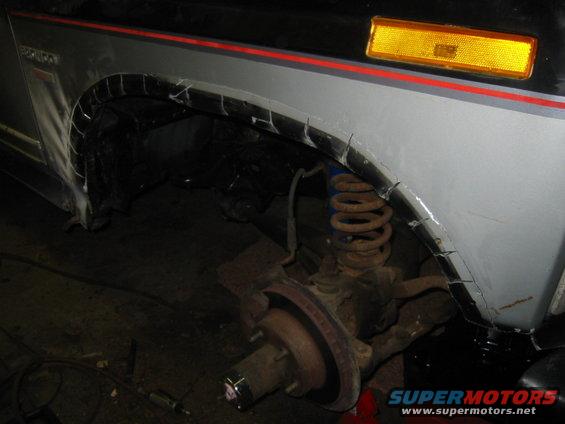 Vehicle's front wheel well with cut fenders and exposed suspension. Gray body with red stripe