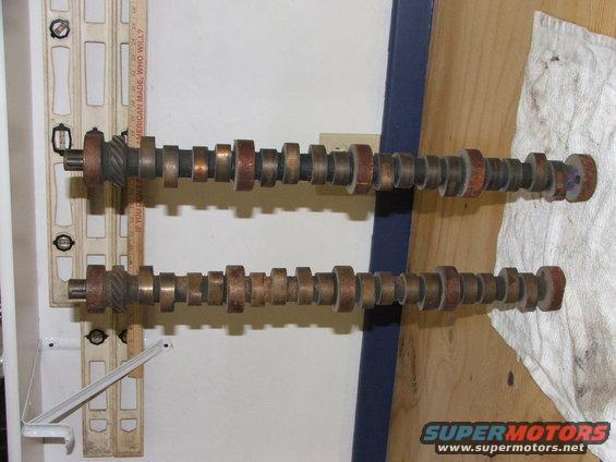 Two camshafts with multiple lobes, one above the other, resting on a wooden surface