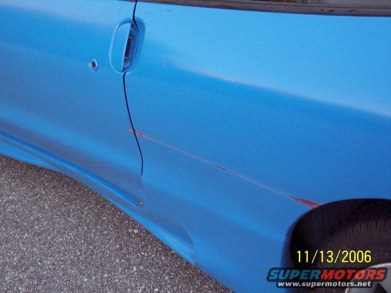 Close-up of a blue car's side door showing a scratch and a door handle. A tire is partially visible