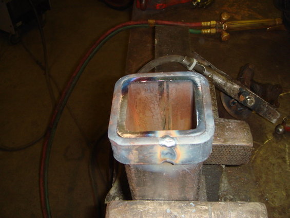 Welding torch with red and green hoses aimed at a metal part held in a vise