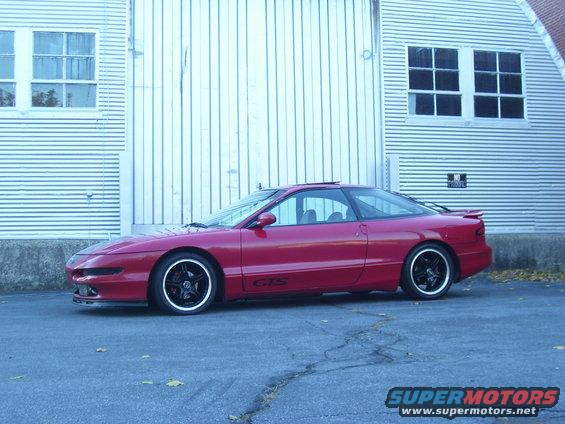 Red 1997 Ford Probe coupe with black rims parked in front of a corrugated metal building