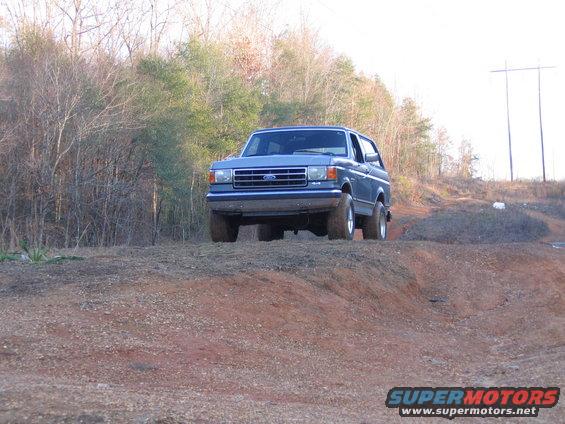 Blue Ford Bronco driving on a dirt hill, front 3/4 view. The vehicle has a blue grille and chrome bumper