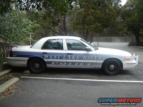 Side view of a white West Lake Hills police car parked in a lot. It has blue stripes and text