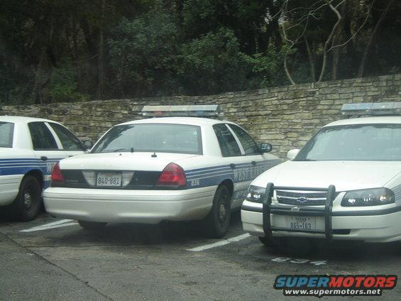 Three white police cars parked, two in full view showing rear and front, one partially visible. The car on the left has a blue stripe and license plate 840-881. The car on the right has a push bar and license plate 865-163