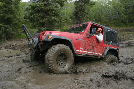 Red Jeep Wrangler stuck in deep mud, driver wearing orange hat visible