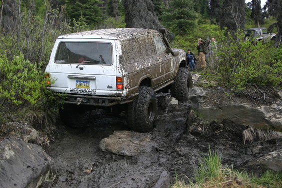 White SUV with snorkel and off-road tires navigating a muddy trail, rear view