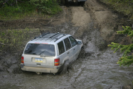 Silver SUV with roof rack stuck in mud, another vehicle visible on the hill above