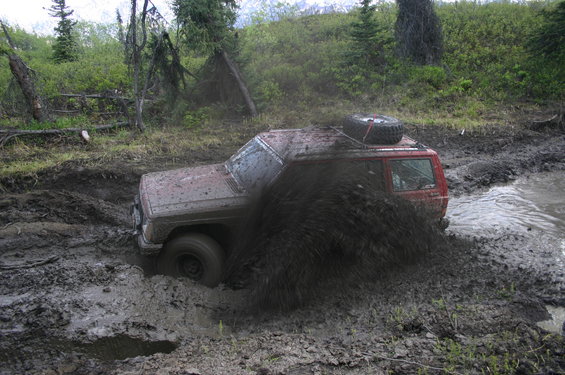 Red SUV with spare tire on roof stuck deep in mud, throwing up a large spray