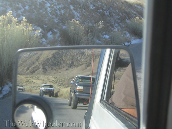 Reflection in side mirror shows three SUVs driving on a road with snowy hills in background