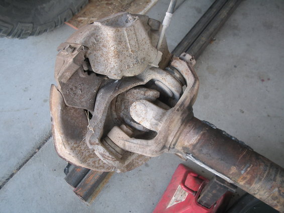 Close-up of a rusty front axle assembly with brake components attached. Some text is visible on a metal plate