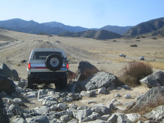 White SUV with spare tire on rear bumper navigates rocky terrain; two other vehicles visible in distance