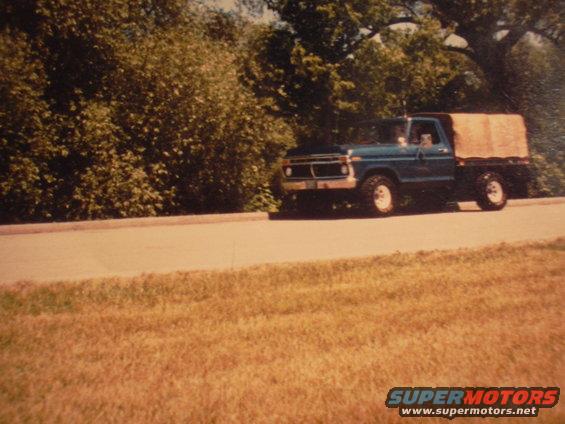 1977 Ford F-150 flatbed truck driving on a road, blue cab with a tarped bed
