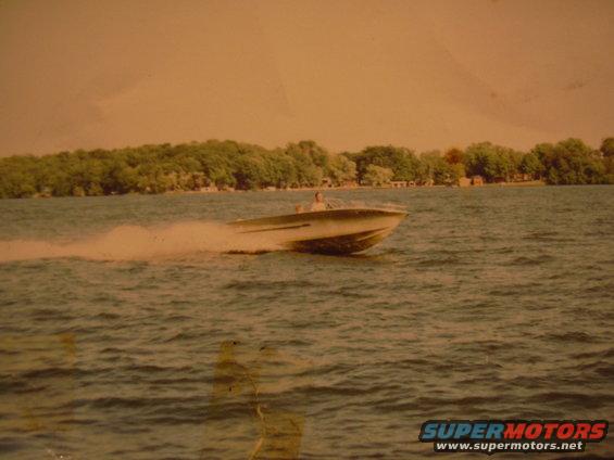 Speed boat cutting through water with a person at the helm. Trees and buildings line the distant shore