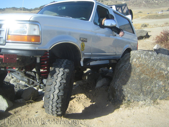 Silver Ford Bronco on rocks, front passenger side tire angled up, driver's arm out window