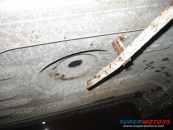 Underside of a vehicle showing a circular indentation and a rusty metal strap