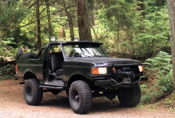 Dark green 1992 Ford Bronco with off-road tires and a winch on the front bumper, parked on a dirt road in a forest