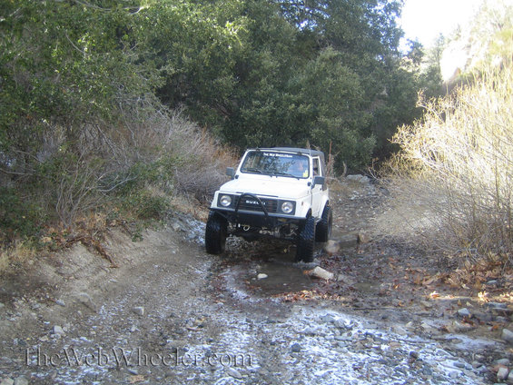 1987 Suzuki Samurai driving through a shallow stream on a dirt trail with trees on either side