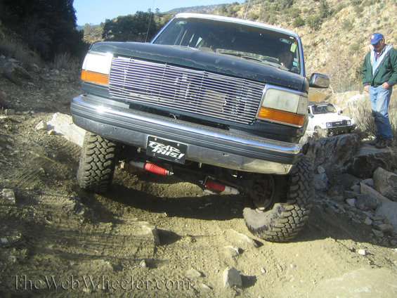 Dark green Ford F-150 truck with chrome grille, lifted suspension, and off-road tires navigating rocky terrain