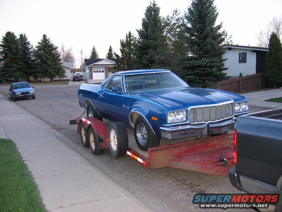 Blue 1976 Ford Ranchero on a red trailer, front 3/4 view, being towed by a dark pickup truck