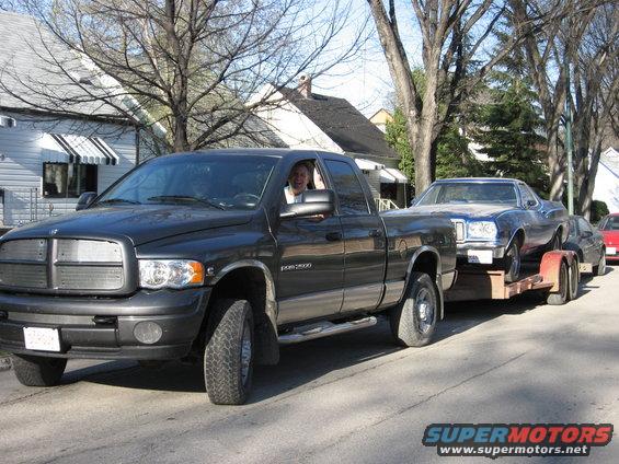 Black Dodge Ram pickup towing a blue car on a trailer down a residential street