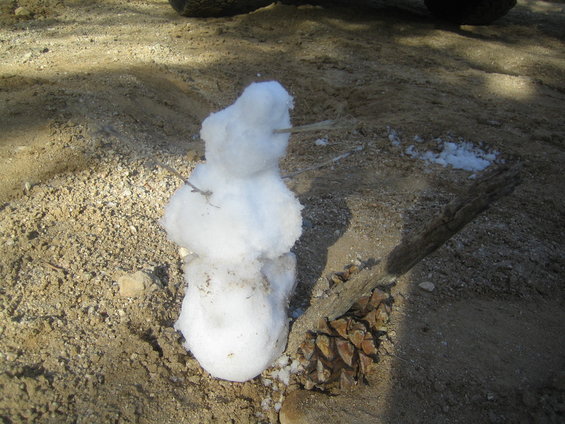 Small snowman made of snow on sandy ground with a pine cone and stick