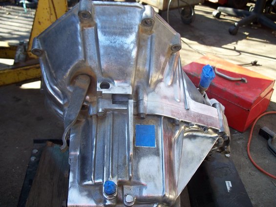 Shiny metal transmission casing with blue caps on fittings. Red toolbox and yellow equipment in background