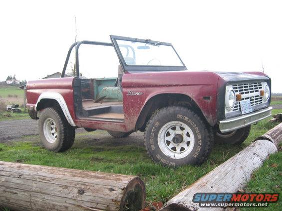 Red 1968 Ford Bronco with doors removed, parked on grass with logs in foreground