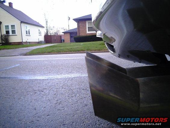 Close-up of a car's rear bumper and exhaust pipe. Houses and a street are visible in the background