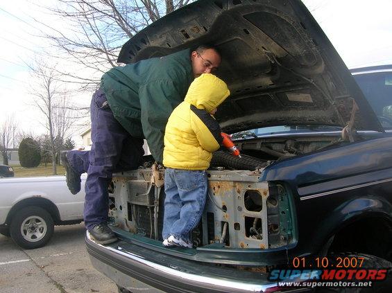 Dark green truck with hood open, father and child inspect engine. White truck visible to the left