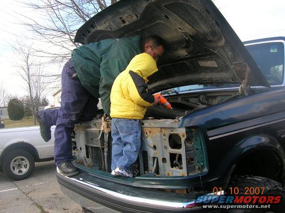 Man and child look at engine bay of dark green truck. White truck in background