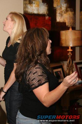 Two women stand indoors, one with blonde hair, the other with brown hair. A lamp and abstract art are visible