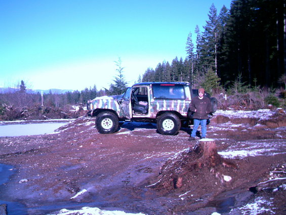 1987 Ford Bronco II with camouflage paint parked on dirt with snow patches, person standing on a tree stump nearby