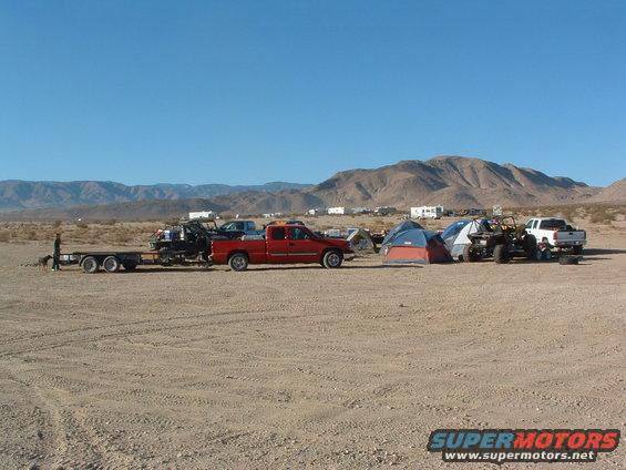 Desert campsite with red pickup truck, trailer, off-road vehicle, white pickup truck, and tents