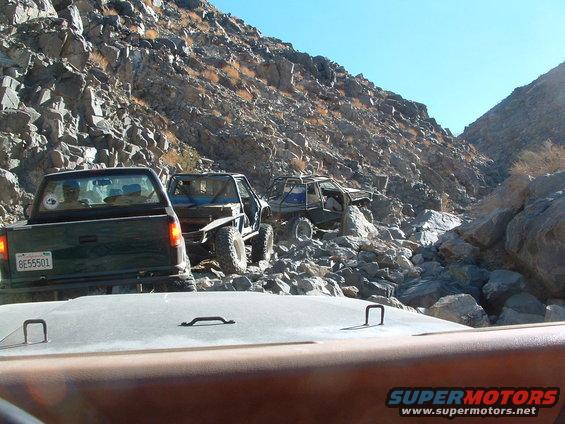 Three off-road vehicles driving through a rocky canyon. The lead vehicle is a dark green pickup truck with license plate 8E55501. Two modified SUVs follow behind