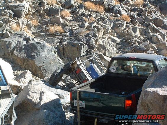 Off-road buggy tipped on its side amidst rocks, with a dark green pickup truck behind it