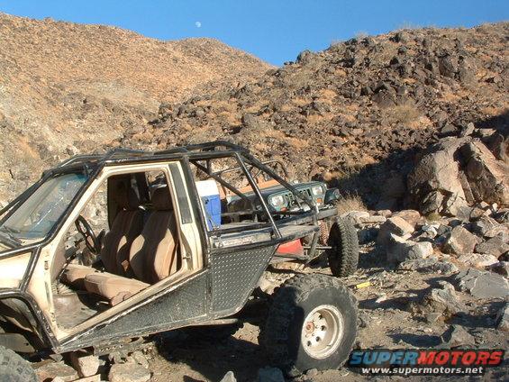 Off-road buggy with roll cage and tan interior, parked next to a green Jeep in a rocky, desert landscape