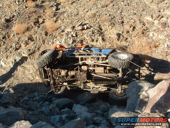 off-road vehicle flipped on its side in rocky terrain with two people looking on