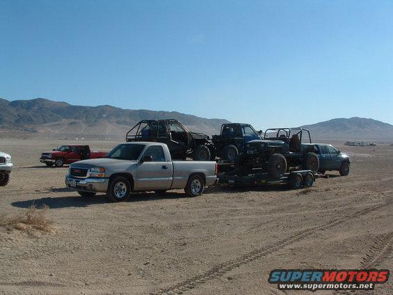Silver GMC pickup towing a trailer with two off-road vehicles. Red pickup and another truck in background