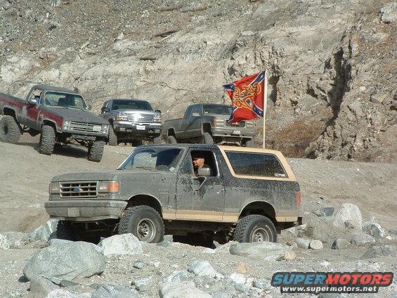 Ford Bronco off-roading on rocky terrain, with three pickup trucks behind it. A Confederate flag is visible
