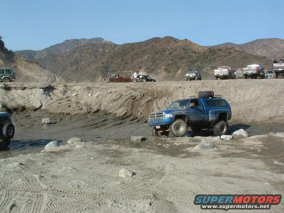 Blue pickup truck with a camper shell driving through a sandy, rocky terrain with mountains in the background. Other vehicles are visible on a ridge