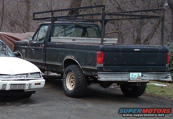 Black Ford pickup truck, rear view, with a ladder rack and toolbox. Visible rust on the body