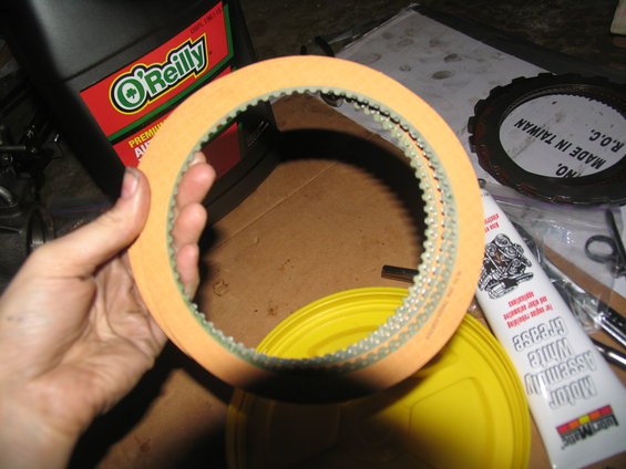 Hand holding orange transmission clutch plates with green teeth. O'Reilly oil bottle and tube of motor assembly grease visible