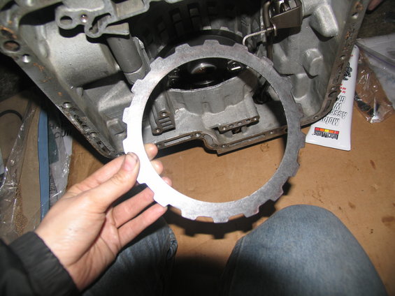 Hand holding a transmission clutch plate in front of an open transmission case. Tube of transmission assembly lube is visible