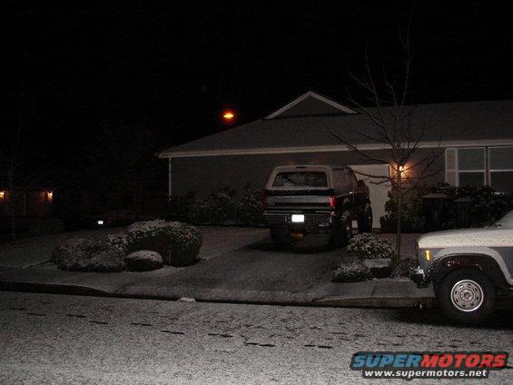 Rear view of a lifted Ford Bronco parked in a driveway at night with snow on the ground. Another vehicle is partially visible in the foreground