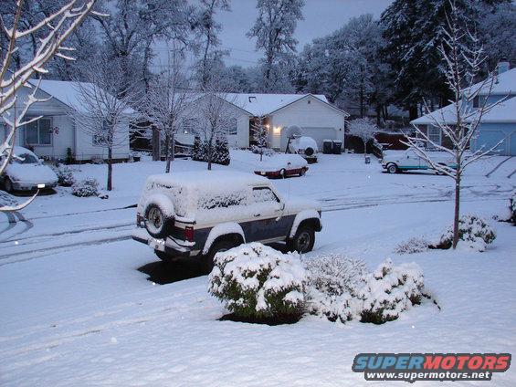 Snow-covered Ford Bronco parked in a driveway with a spare tire on the back. Other cars and houses are visible in the background
